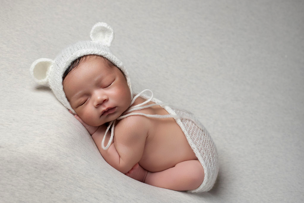 A newborn boy in a knit bear-ears bonnet, sleeping on a textured cream blanket inside the Studio Realms studio.