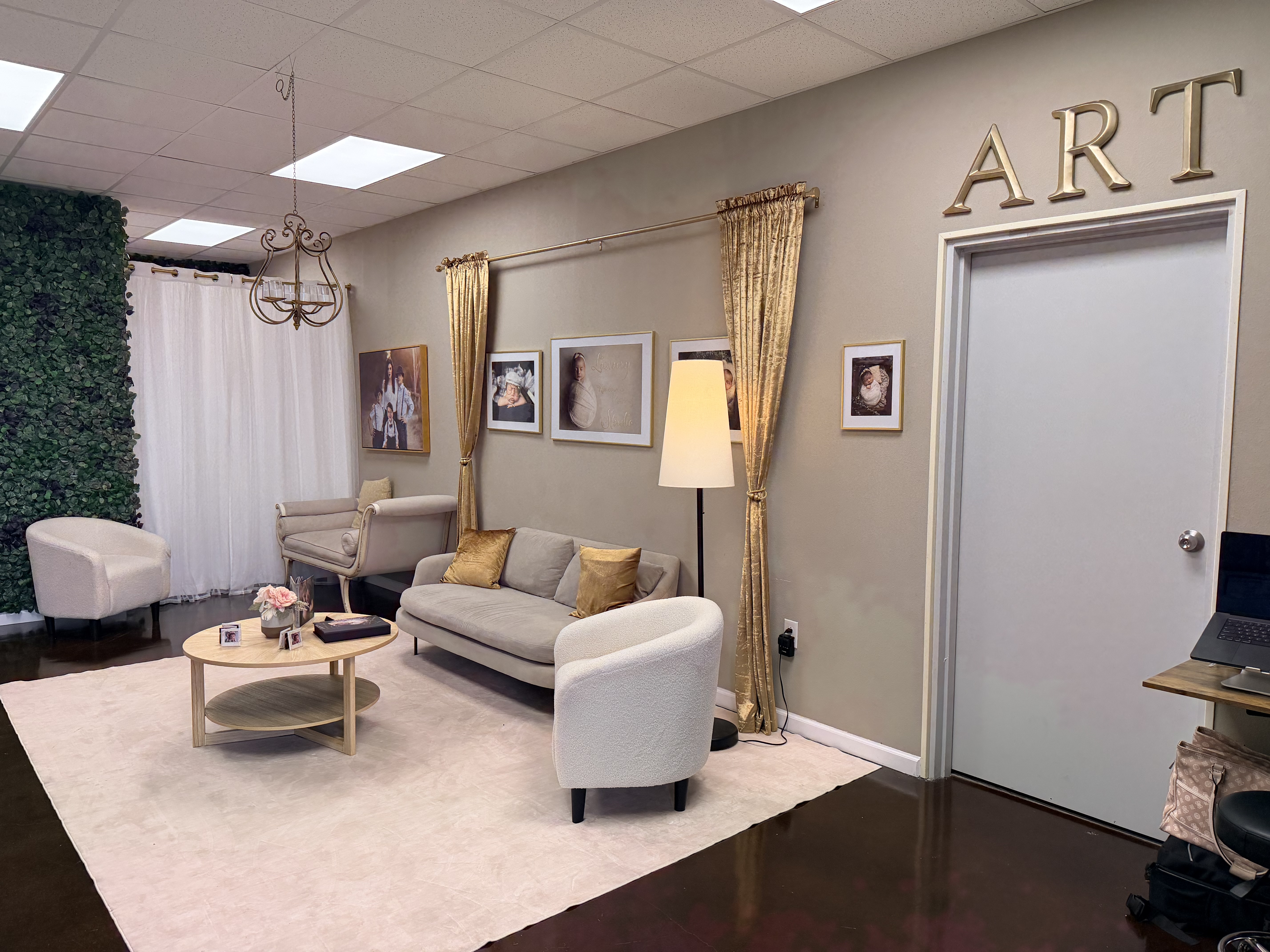 The Studio Realms client lounge: a soft sage couch with gold accent pillow, gold-framed newborn portrait prints on warm gray walls, gold curtains, and an ART feature wall.